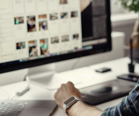 A person uses a desktop computer displaying photo thumbnails, while exploring web design ideas and a color swatch on a nearby laptop. Wearing a smartwatch, they work in marketing at a modern desk workspace. {{brizy_dc_image_alt imageSrc=