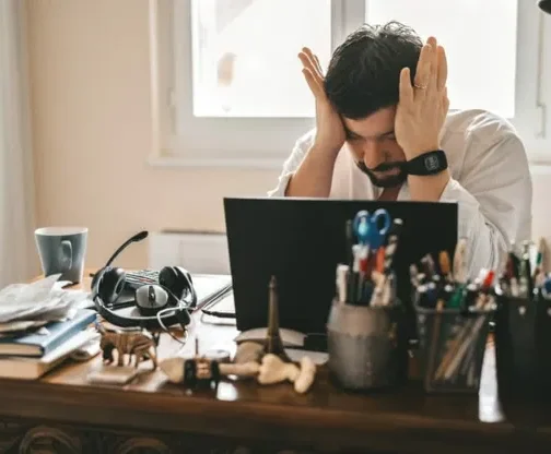 A man sits at a cluttered desk with his hands on his head, looking stressed while working on SEO marketing tasks on his laptop. The desk has books, a headset, pens, small figurines, and a window in the background. {{brizy_dc_image_alt entityId=